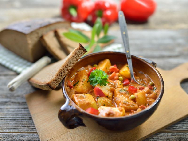 Getöpferte Schale mit Kartoffel-Gulasch auf einem Brett und drei Scheiben Brot daneben.