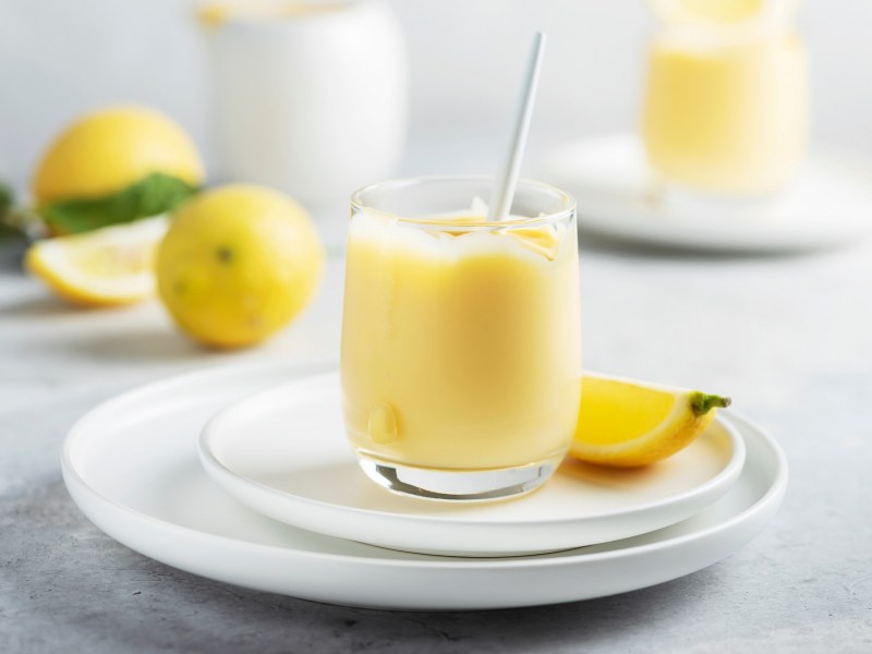 Ein Glas Limoncello-Creme auf einem Teller, darauf auch eine Zitronenspalte, im Hintergrund unscharf zwei weitere Gläser und Zitronen.