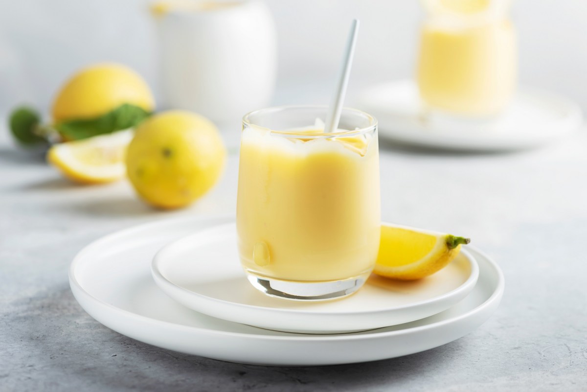 Ein Glas Limoncello-Creme auf einem Teller, darauf auch eine Zitronenspalte, im Hintergrund unscharf zwei weitere Gläser und Zitronen.