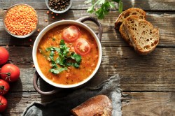 Eine Schüssel mit Henkeln, darin Linsen-Tomaten-Suppe, garniert mit halbierten Tomaten und Koriander, daneben frische Tomaten, Brotscheiben, eine Schale Linsen, Draufsicht.