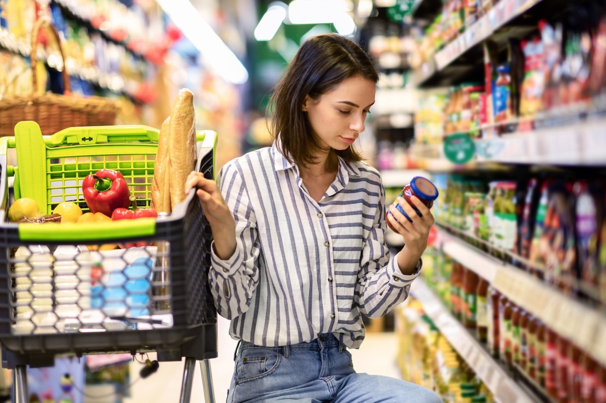 Eine dunkelhaarige junge Frau prüft im Supermarkt das Mindesthaltbarkeitsdatum eines Lebensmittels im Glas und hockt dabei neben ihrem Einkaufswagen.