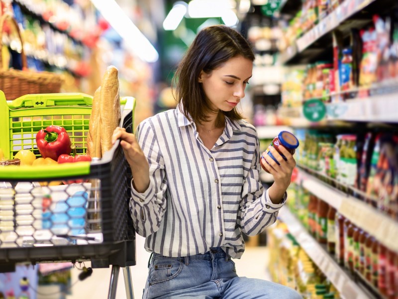 Eine dunkelhaarige junge Frau prüft im Supermarkt das Mindesthaltbarkeitsdatum eines Lebensmittels im Glas und hockt dabei neben ihrem Einkaufswagen.