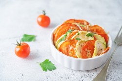 Mini-Gemüse-Gratin mit Tomate und Zucchini in einer weißen runden Auflaufform, daneben Mini-Tomaten.