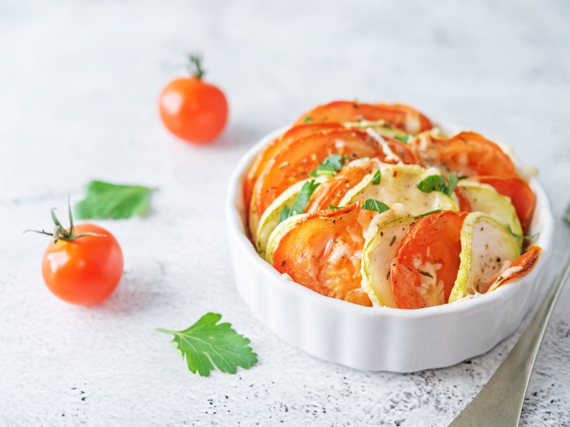 Mini-Gemüse-Gratin mit Tomate und Zucchini in einer weißen runden Auflaufform, daneben Mini-Tomaten.
