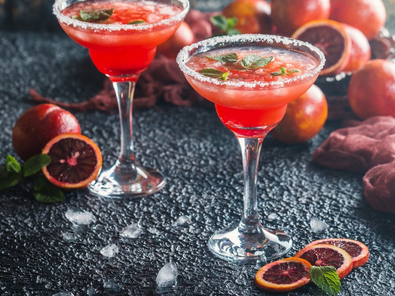 Zwei Gläser mit rotem Cocktail und Blutorangen auf einem Tisch.