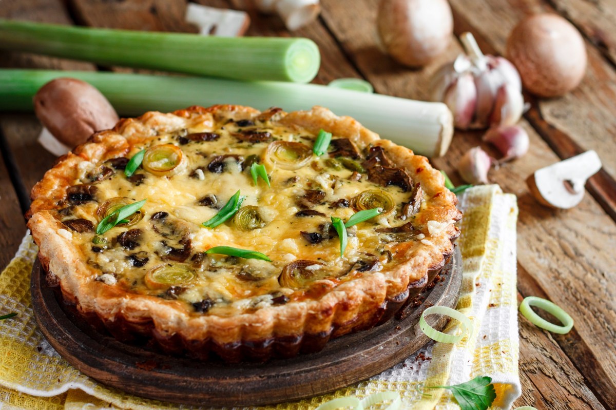 Eine Pilz-Lauch-Quiche, daneben ganze Pilze und Lauch auf Holzuntergrund.