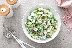 Schale mit Salat mit Radieschen und Gurken auf einem hellen Tisch und Besteck.