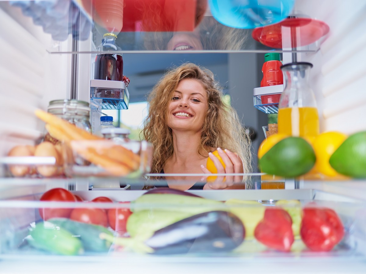 Salz im Kühlschrank: Frau mit blonden Locken guckt lachend in den Kühlschrak, der mit viel Gemüse und Obst gefüllt ist.