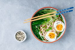 Eine Schüssel Erdnussbutter-Ramen mit halbiertem Ei und Pak Choi in der Draufsicht, darauf Stäbchen, daneben eine Schale Sesam.