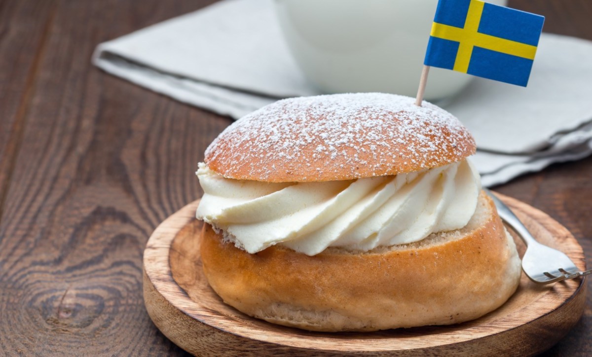 Eine schwedische Semla auf einem Holbrett mir einem Fähnchen der schwedischen Flagge darin. Im hintergrund steht eine Tasse Kaffee.