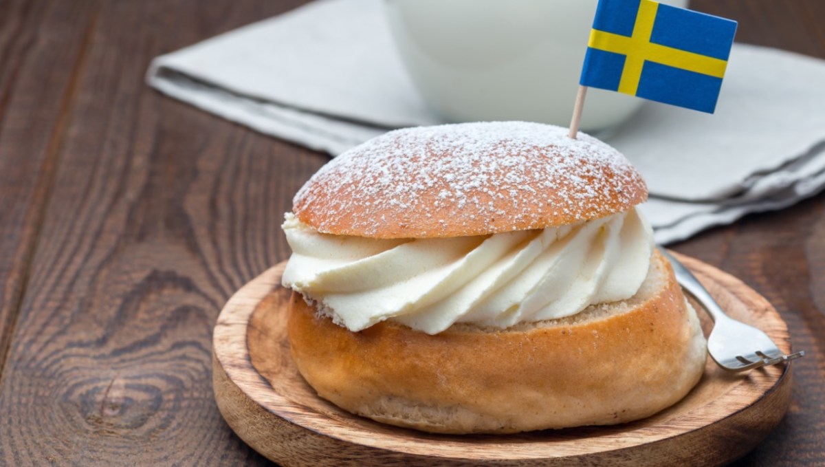 Eine schwedische Semla auf einem Holbrett mir einem Fähnchen der schwedischen Flagge darin. Im hintergrund steht eine Tasse Kaffee.