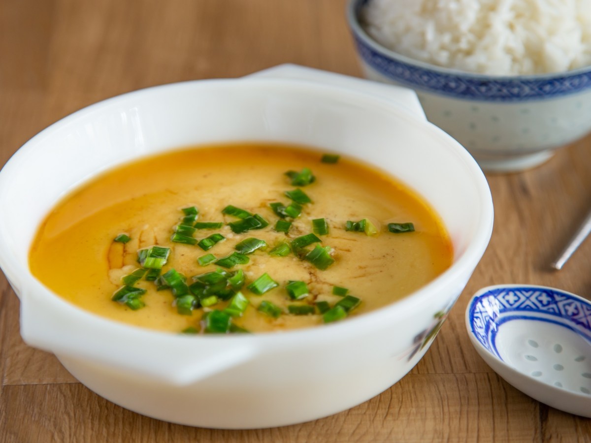 Steamed Eggs in einer Porzellanschale, garniert mit Schnittlauch und Sojoasoße. Daneben eine Schale Reis.