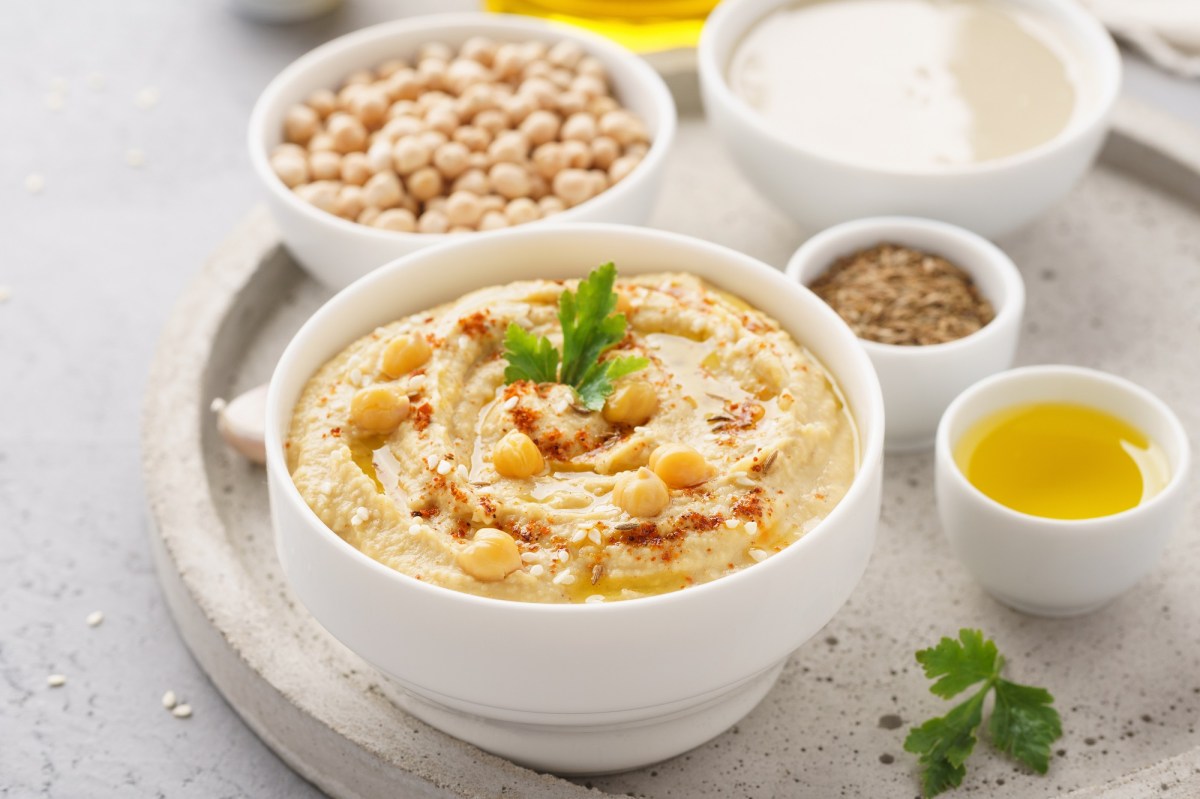 Tipps Hummus: eine weiße Schale Hummus mit Kichererbsen und Koriander sowie Sesam garniert, daneben je eine Schüssel rohe Kichererbsen, Öl, Gewürz und Kichererbsenwasser.
