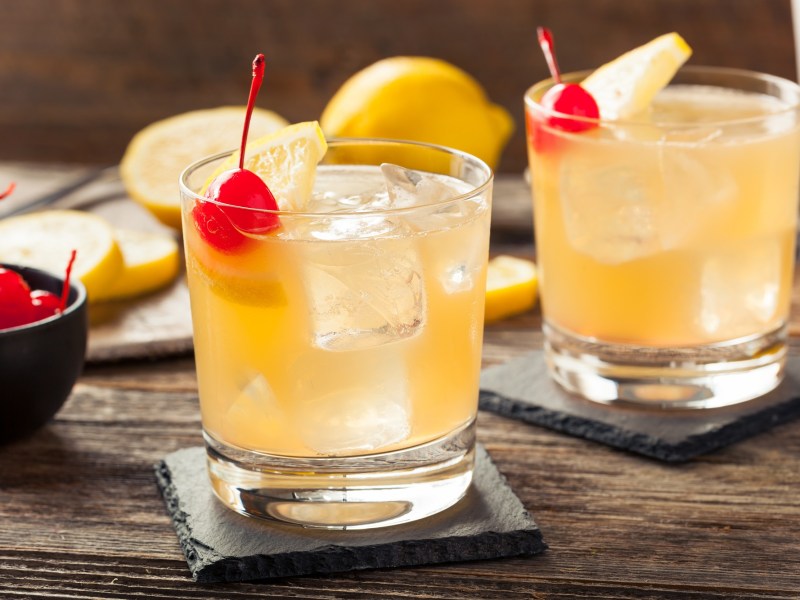 zwei Gläser Whiskey Sour auf Eis mit Cocktailkirschen garniert.