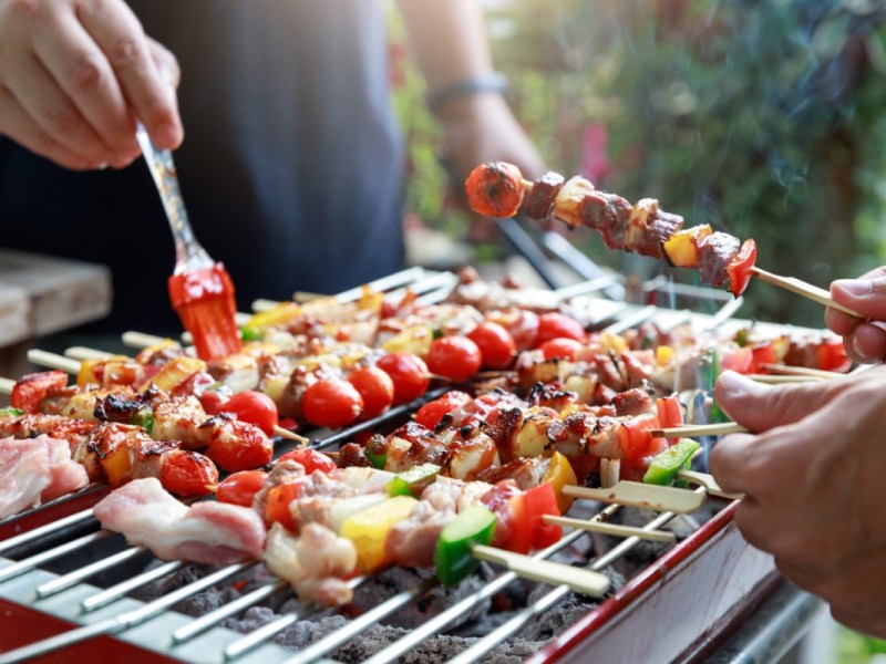 Angrillen: Grillgut auf einem Grill. Zwei Personen stehen um den Grill herum und halten Spieße und einen Pinsel in der Hand.