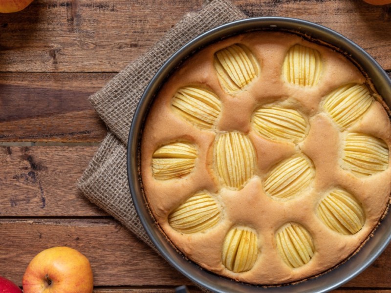 Ein Apfelkuchen ohne Zucker in der Backform von oben fotografiert. Es liegen frische Äpfel daneben.