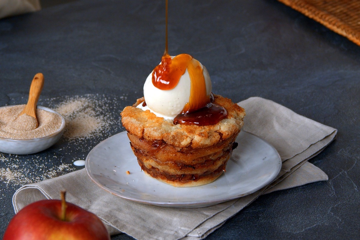 Ein Apfelmuffin ohne Teig mit Vanilleeis auf einem Teller, über den gerade Karamellsoße gegossen wird. Es sind ein Apfel und eine Schale mit Zimtzucker zu sehen.