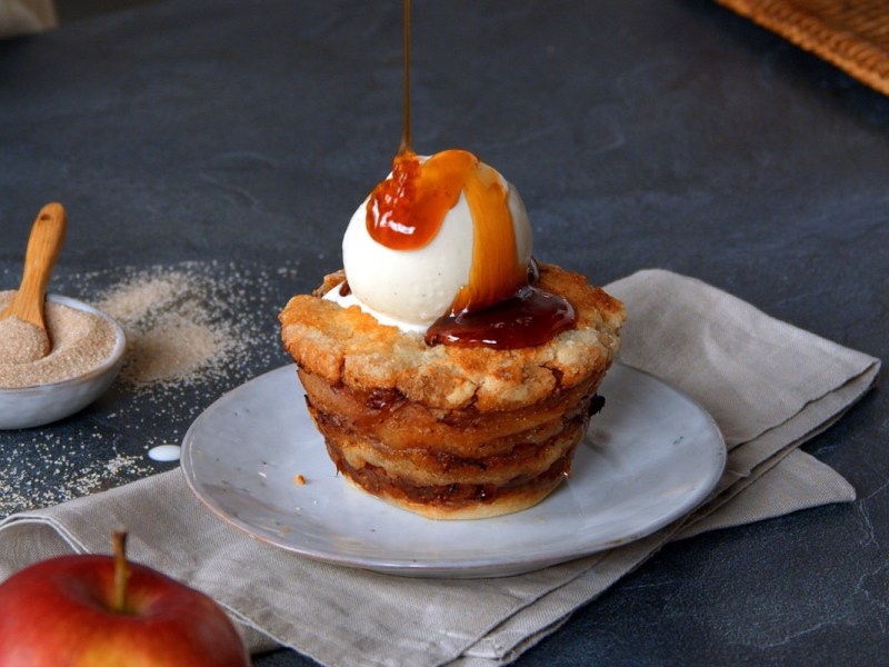 Ein Apfelmuffin ohne Teig mit Vanilleeis auf einem Teller, über den gerade Karamellsoße gegossen wird. Es sind ein Apfel und eine Schale mit Zimtzucker zu sehen.