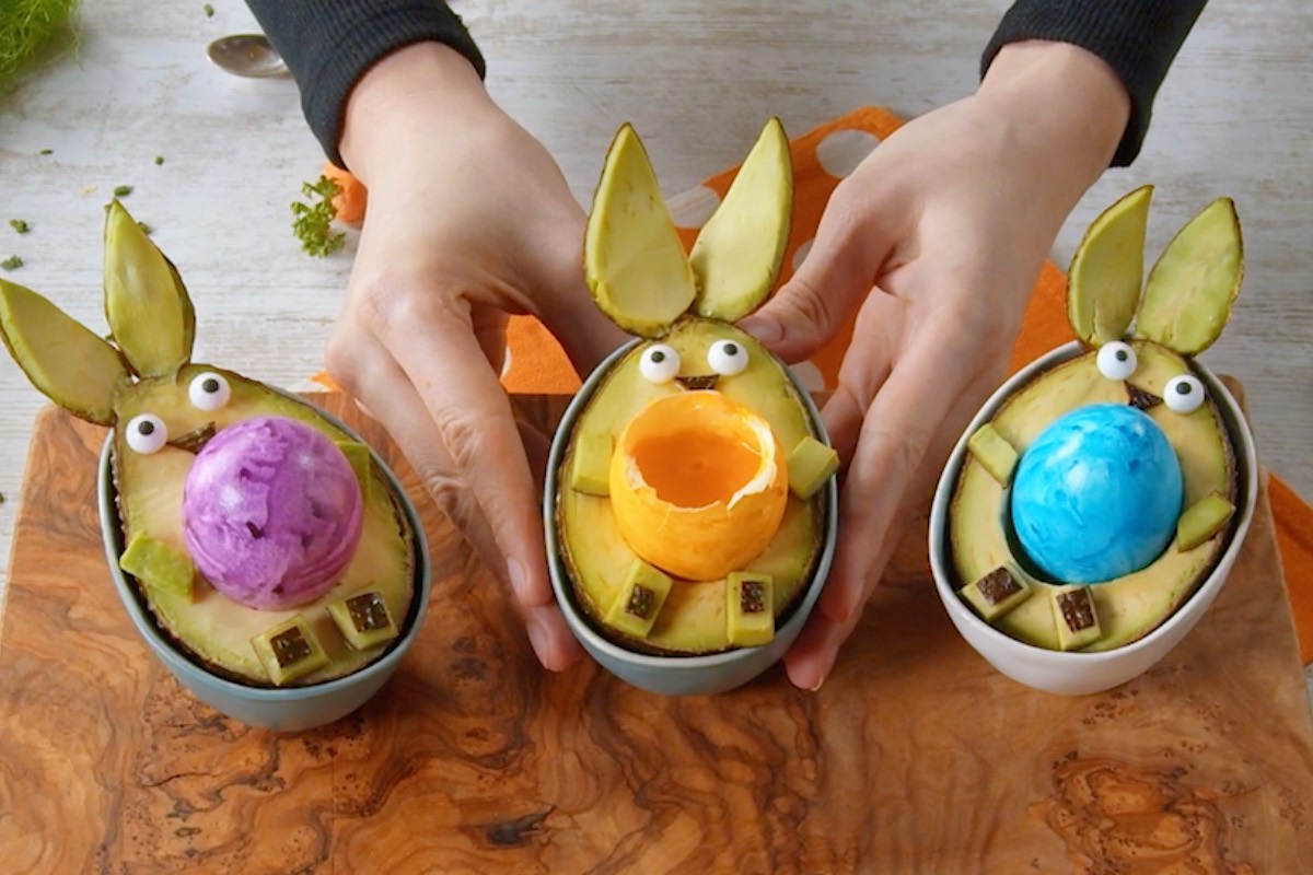 3 Avocado-Hasen mit bunten Eiern. Der mittlere Bunnycado wird von zwei Händen gehalten.