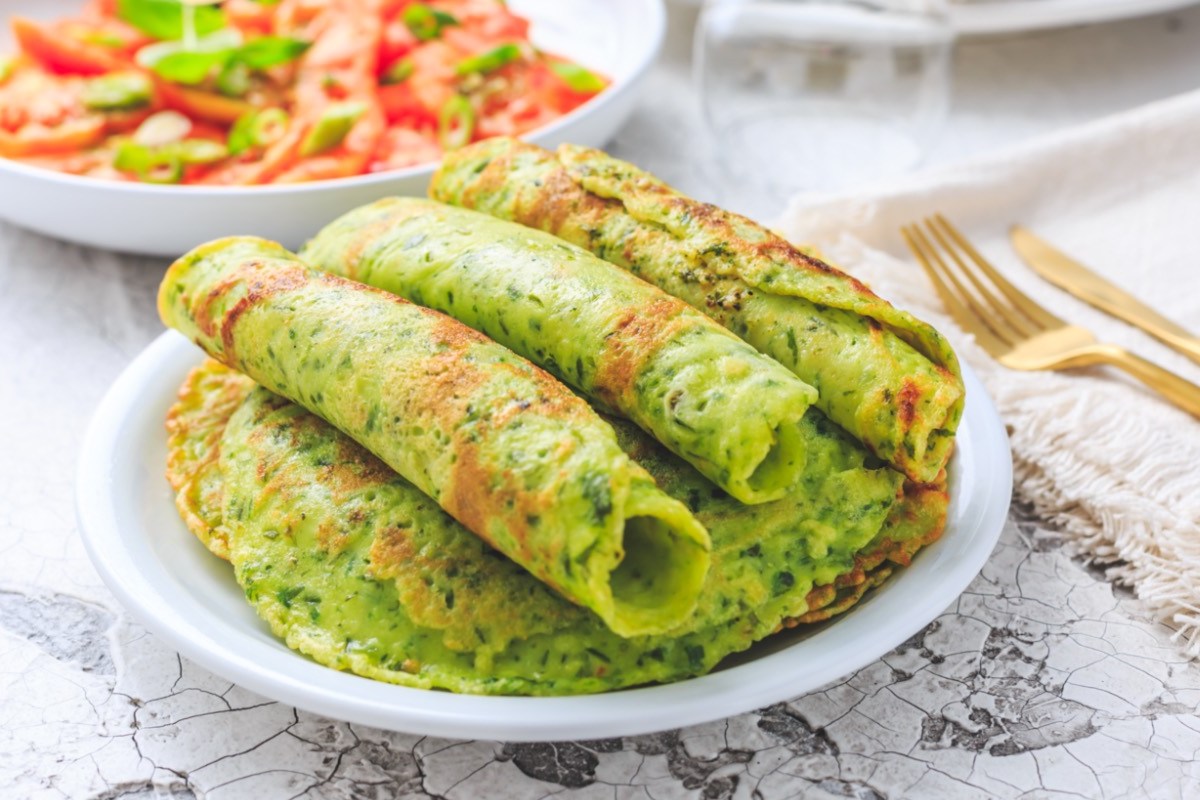 Mehrere gerollte Bärlauch-Pfannkuchen auf einem Stapel weiterer Pfannkuchen. Im Hintergrund ist ein Salat mit Tomaten zu sehen.