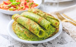 Mehrere gerollte Bärlauch-Pfannkuchen auf einem Stapel weiterer Pfannkuchen. Im Hintergrund ist ein Salat mit Tomaten zu sehen.