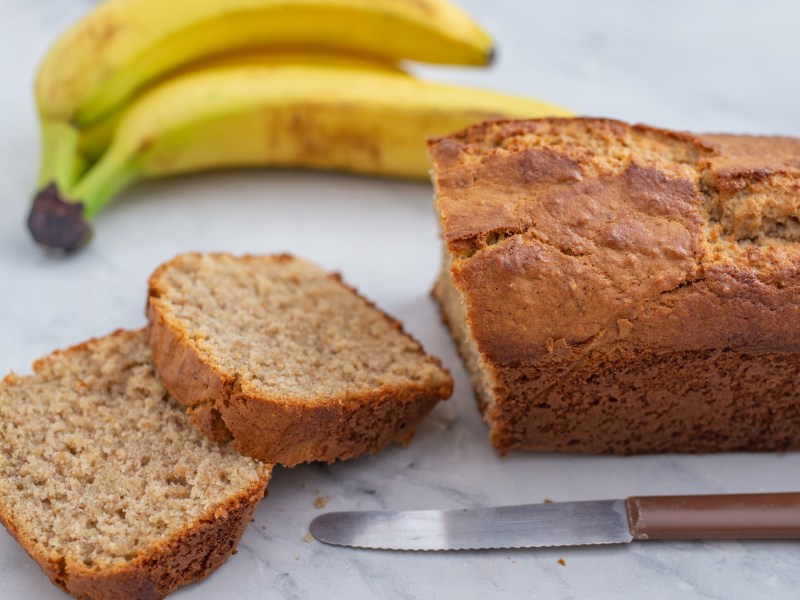 Angeschnittenes Bananenbrot ohne Ei, davor ein Messer, dahinter zwei Bananen.