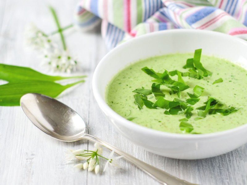 Schüssel mit Bärlauchsuppe, Löffel und Bärlauchblätter auf hellem Tisch