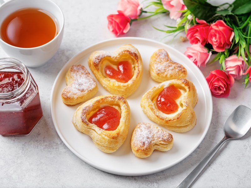 Teller mit Blätterteig-Herzen mit Marmelade, Tee und Rosen auf hellem Untergrund.