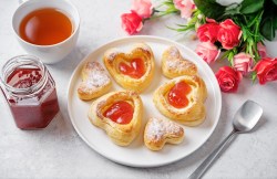 Teller mit Blätterteig-Herzen mit Marmelade, Tee und Rosen auf hellem Untergrund.
