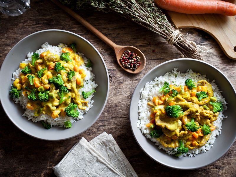 Brokkoli-Curry mit Linsen, angerichtet in zwei Schalen mit Reis. Daneben liegt Besteck als Deko.