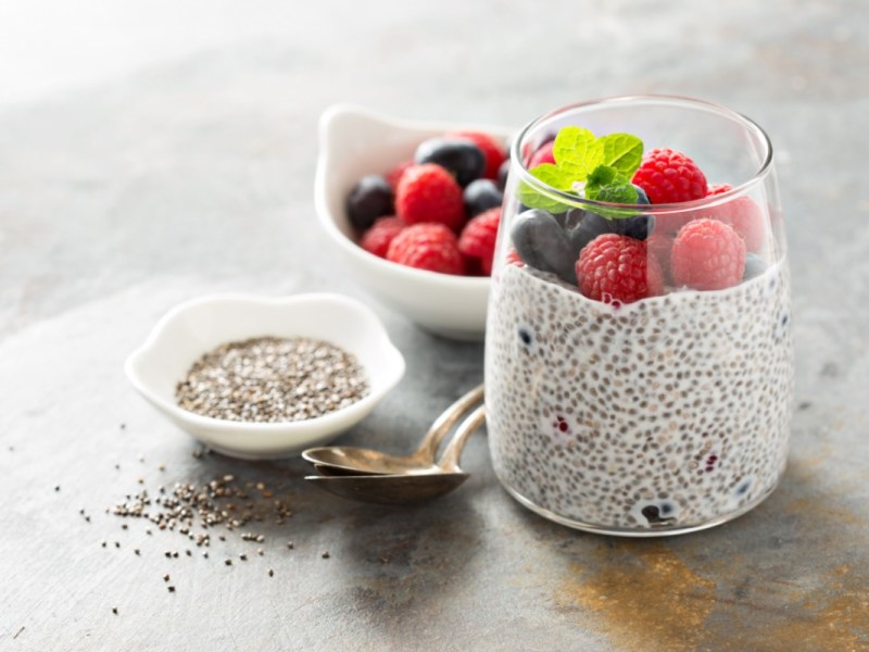 Ein Glas Chia-Pudding mit Himbeeren und Blaubeeren. Daneben stehen eine Schale mit Chiasamen und eine mit Beeren.