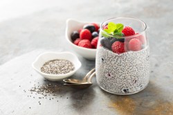 Ein Glas Chia-Pudding mit Himbeeren und Blaubeeren. Daneben stehen eine Schale mit Chiasamen und eine mit Beeren.