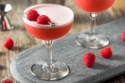 Ein Clover Club Cocktail mit 2 Himbeeren in einem langstieligen Cocktailglas. Himbeeren und ein weiteres Glas sind im Hintergrund zu sehen.