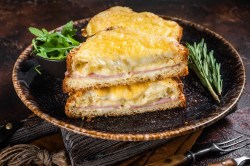 Zwei diagonal durchgeschnittene Croque Monsieur auf einem dunklen Teller. Daneben liegen ein paar Zweige Rosmarin sowie eine Schale Rucola.