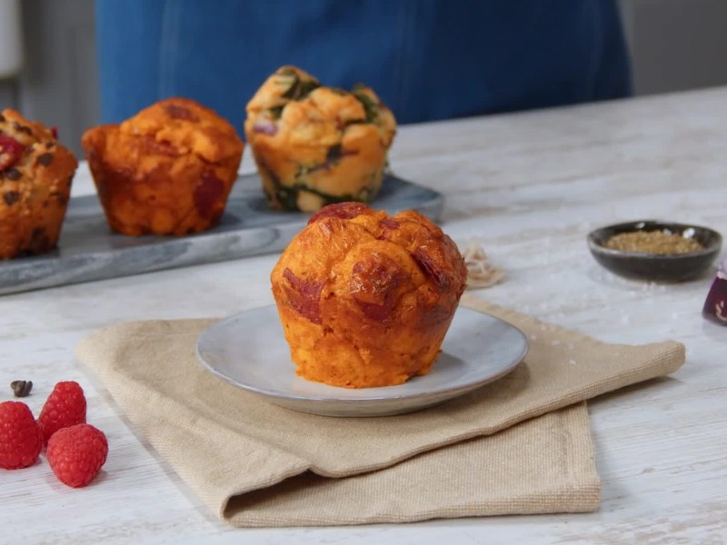 Dreierlei Brötchenmuffins auf einem langen Teller, davor ein einzelner Käse-Salami-Muffin auf einem kleineren Teller. Dieser steht auf einer beigen Serviette. Daneben liegen Himbeeren.