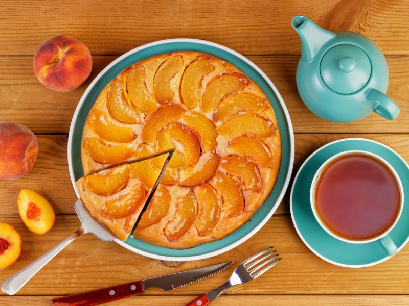Einfacher Pfirsichkuchen, serviert auf einem hübschen blauen Teller. Daneben stehen eine blaue Kaffeetasse und eine Teekanne. Als Dekoration liegen Pfirsiche daneben.