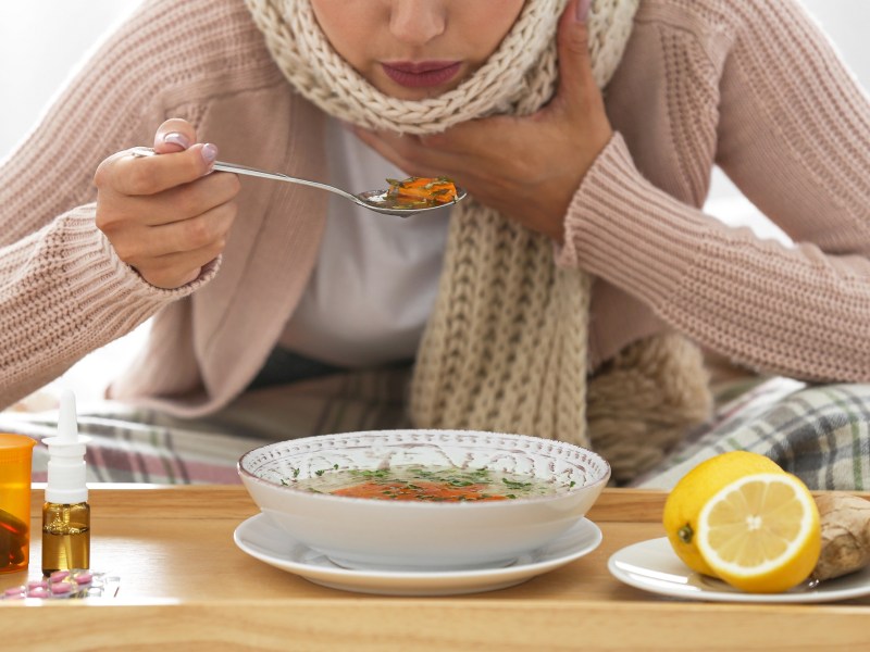 Eine Frau mit Schal isst eine Suppe, ein gutes Essen bei Erkältung. Neben ihr liegen Ingwer, Zitronen und Medikamente.