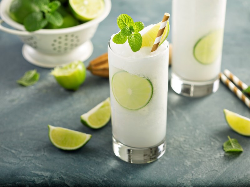 2 Gläser Frozen Gin Tonic mit Minze und Limetten und Strohhalm, drumherum Limettenspalten.