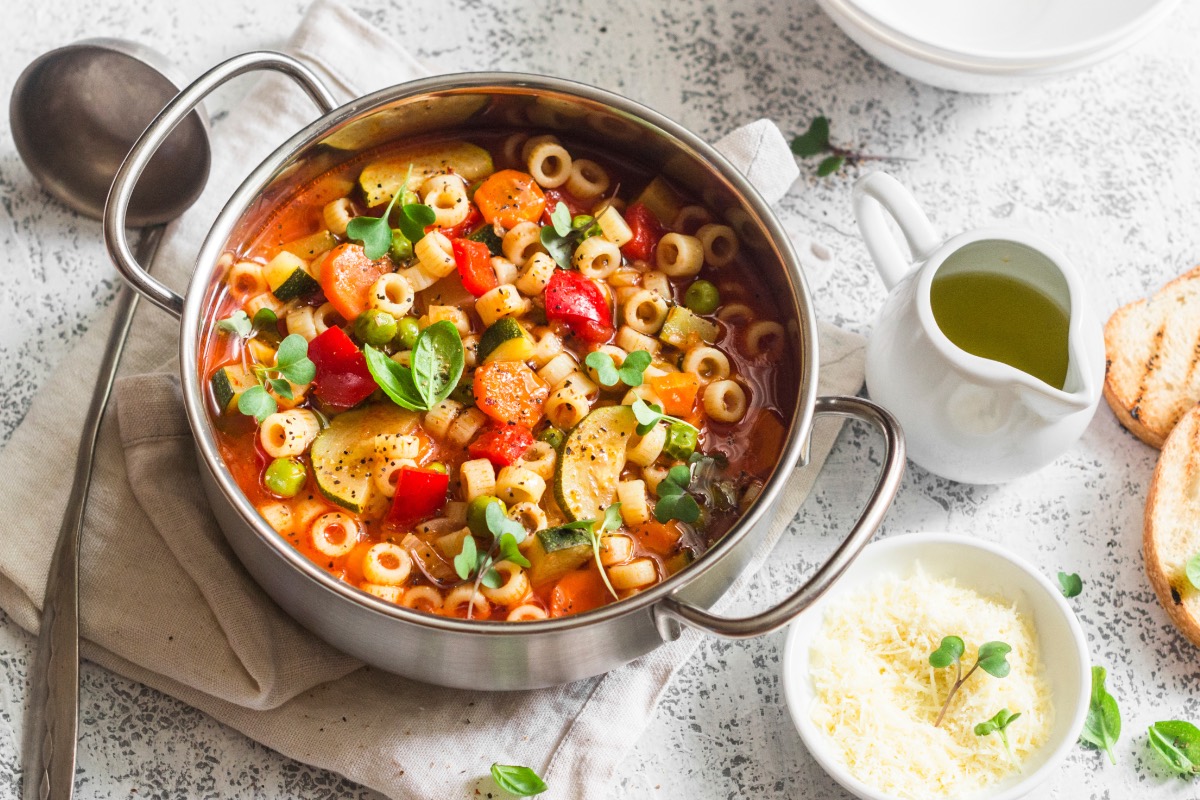 Ein Topf mit Frühlings-Minestrone in der Draufsicht. Daneben liegt eine Kelle und es stehen ein Schälchen mit Käse und eine Kanne mit Öl herum.