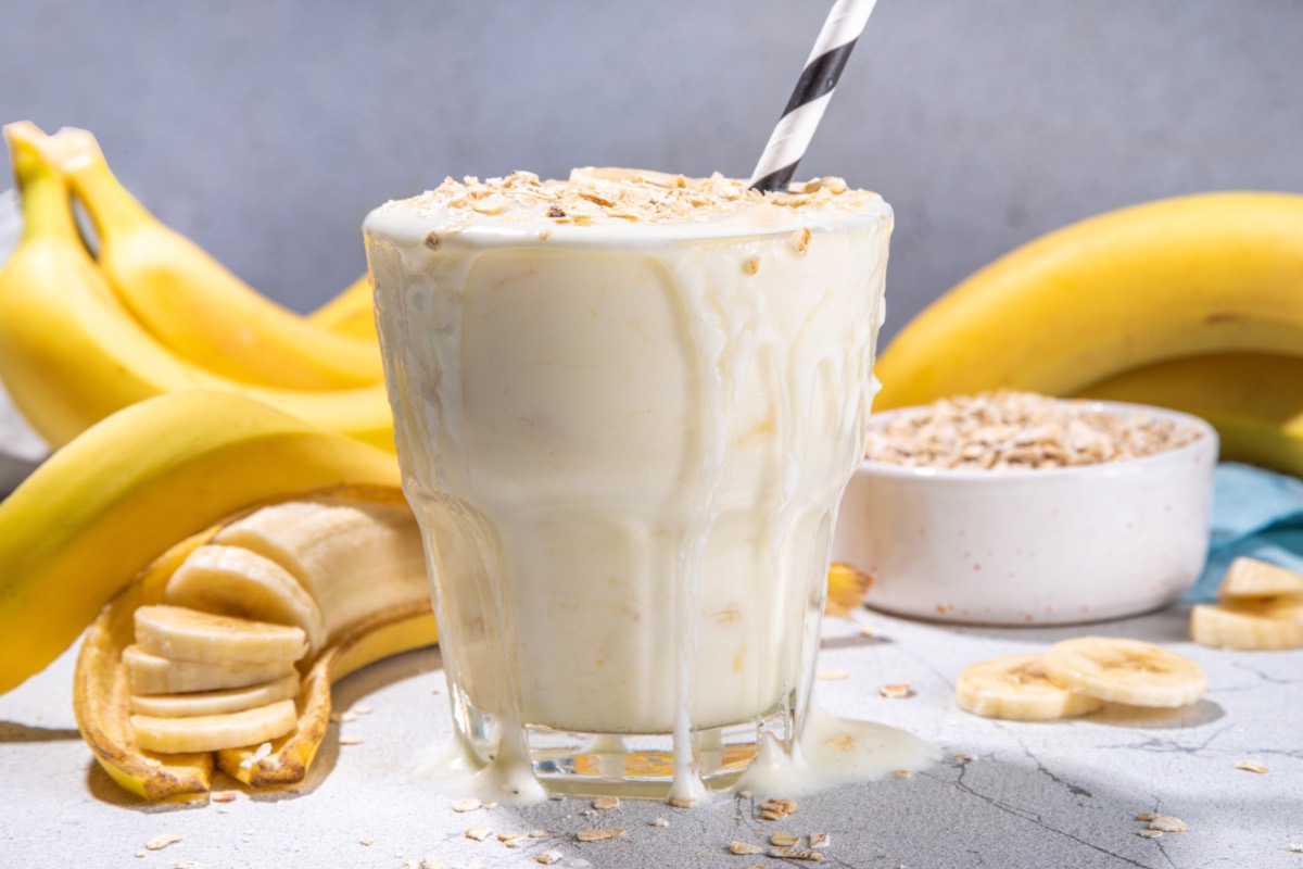 Ein Glas Frühstücksshake mit Bananen und Haferflocken. Im Hintergrund sind Bananen uds eine Schale mit Haferflocke zu sehen.