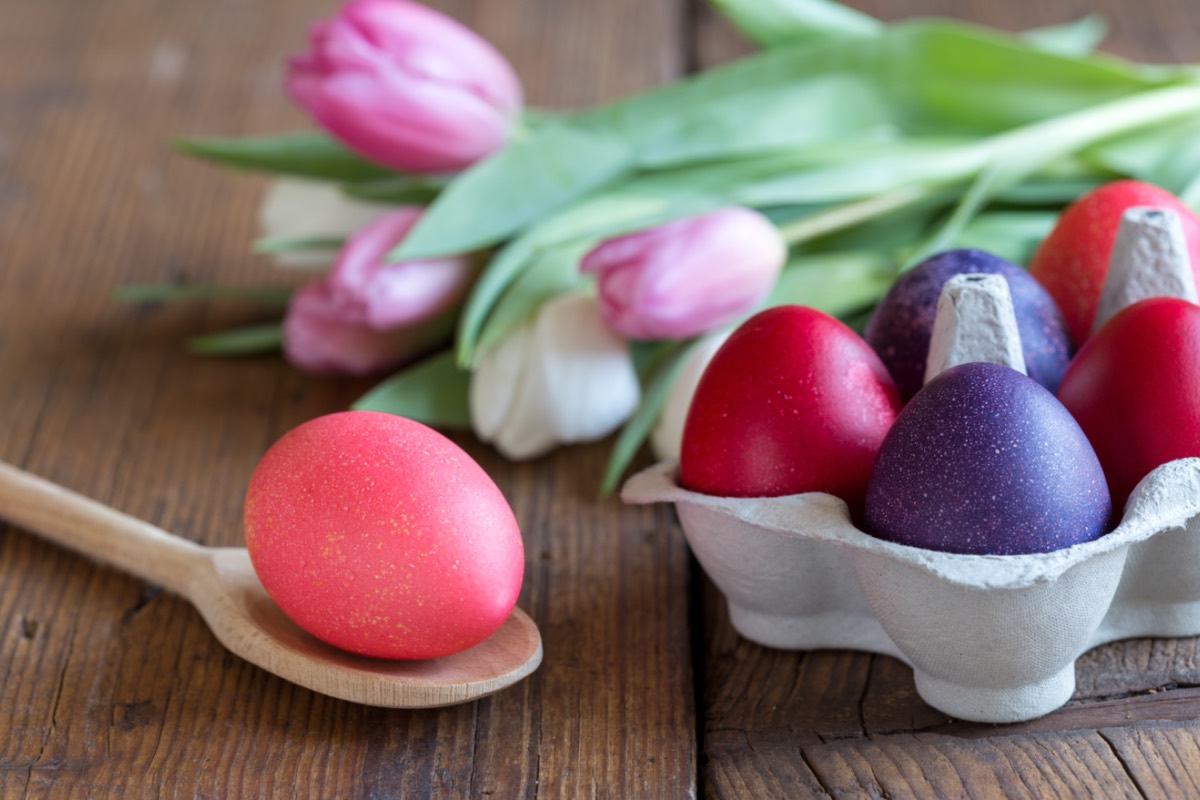 Gefärbte Eier im Eierkarton und auf einem Holzlöffel. Dahinter liegt ein Strauß Tulpen.