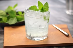 Ein Glas Gin Gin Mule mit Minze auf einem Holzbrett. Im Hintergrund ist frische Minze zu sehen.
