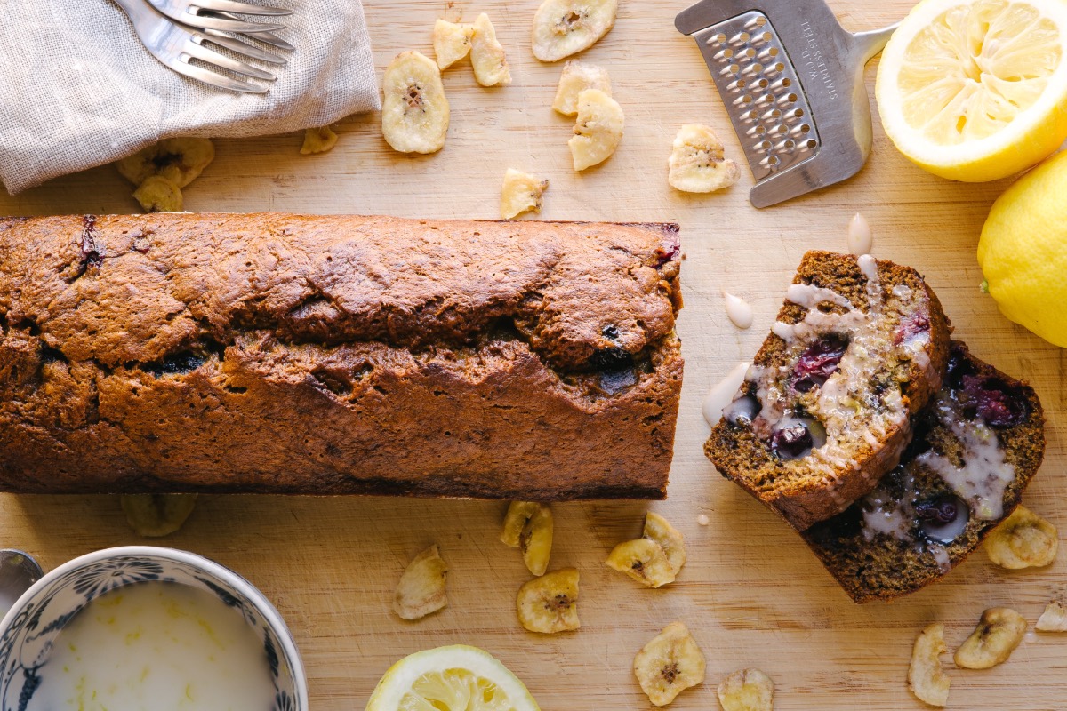 Ein Himbeer-Bananenbrot, von dem ein Stück abgeschnitten wurde in der Draufsicht. Drumherum liegen Küchenwerkzeuge und Zutaten.