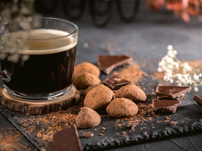 Schieferplatte mit Schokoladenstücken und Kaffee-Pralinen und einem Glas schwarzen Kaffee.