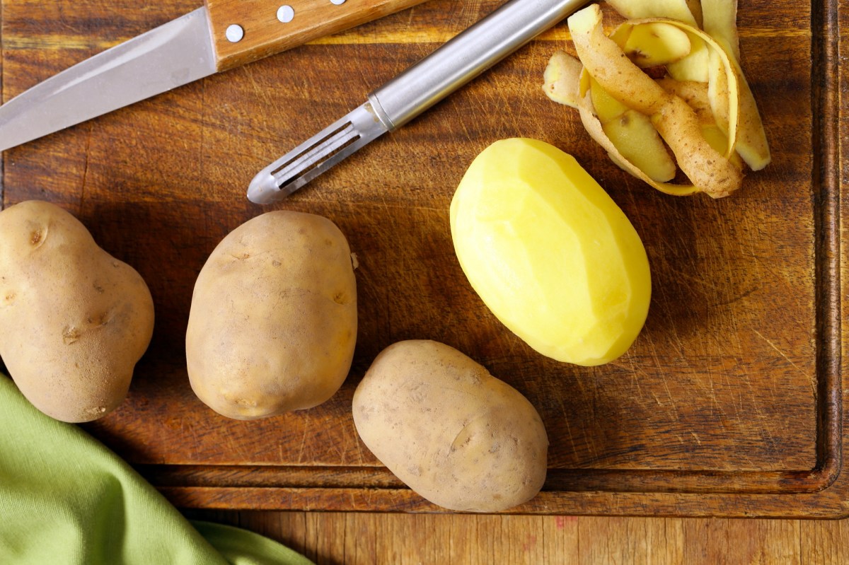 Kartoffel schälen Trick: drei ungeschälte und eine geschälte Kartoffel auf einem Brett, auf dem auch ein Sparschäler und ein Messer sowie Schale liegen.
