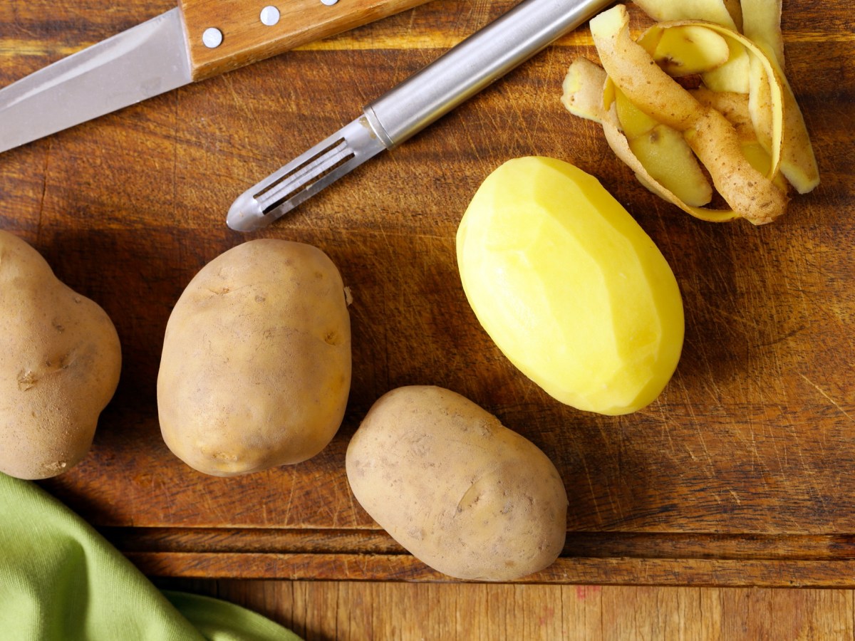 Kartoffel schälen Trick: drei ungeschälte und eine geschälte Kartoffel auf einem Brett, auf dem auch ein Sparschäler und ein Messer sowie Schale liegen.
