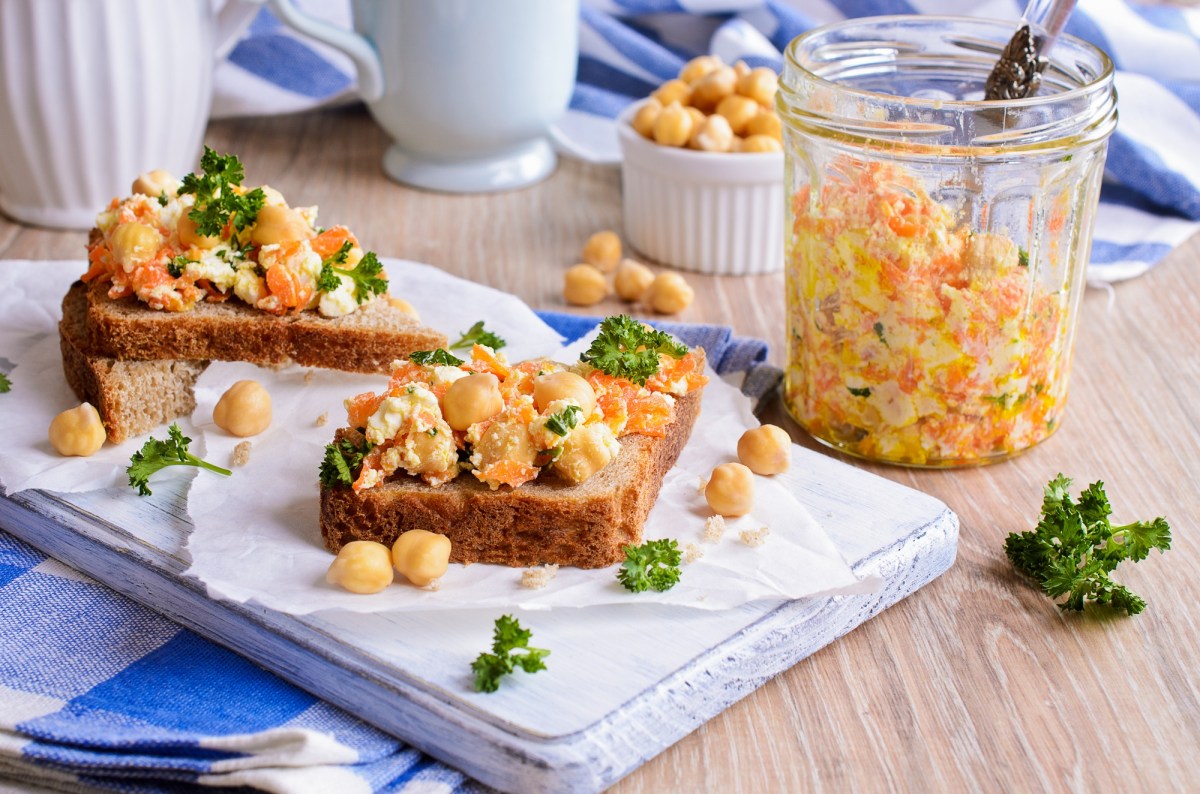 zwei Kichererbsen-Karotten-Stullen auf einem weißen Brett, daneben eine Schale Kichererbsen und ein Glas mit dem Salat.