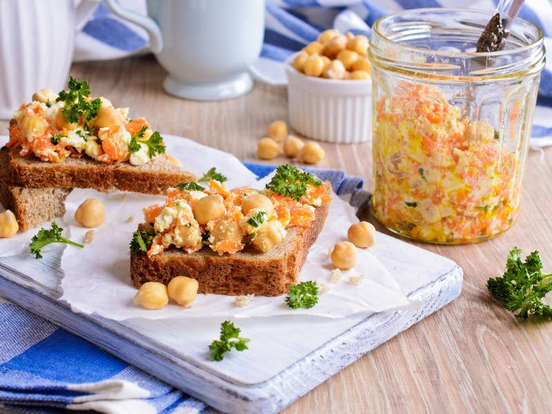 zwei Kichererbsen-Karotten-Stullen auf einem weißen Brett, daneben eine Schale Kichererbsen und ein Glas mit dem Salat.