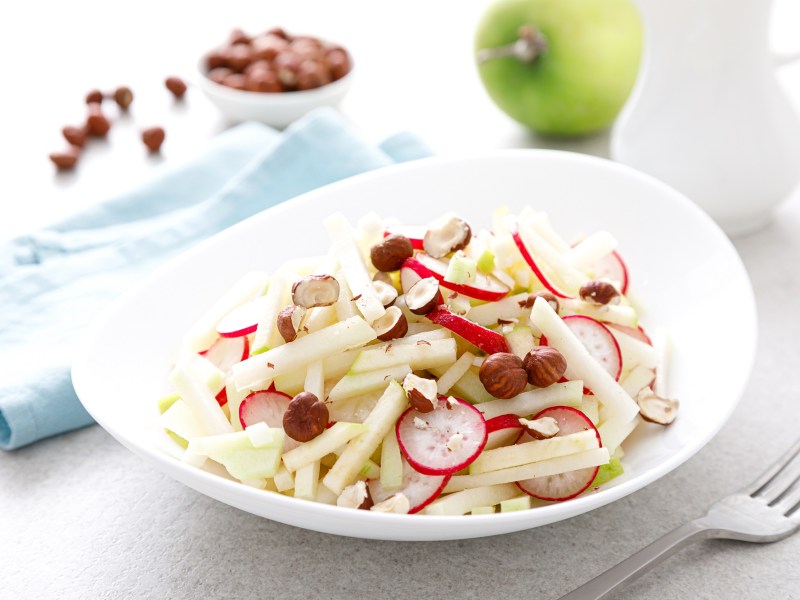 Eine weiße Schale mit Kohlrabi-Apfel-Salat, Nüsse und Apfel auf hellem Tisch