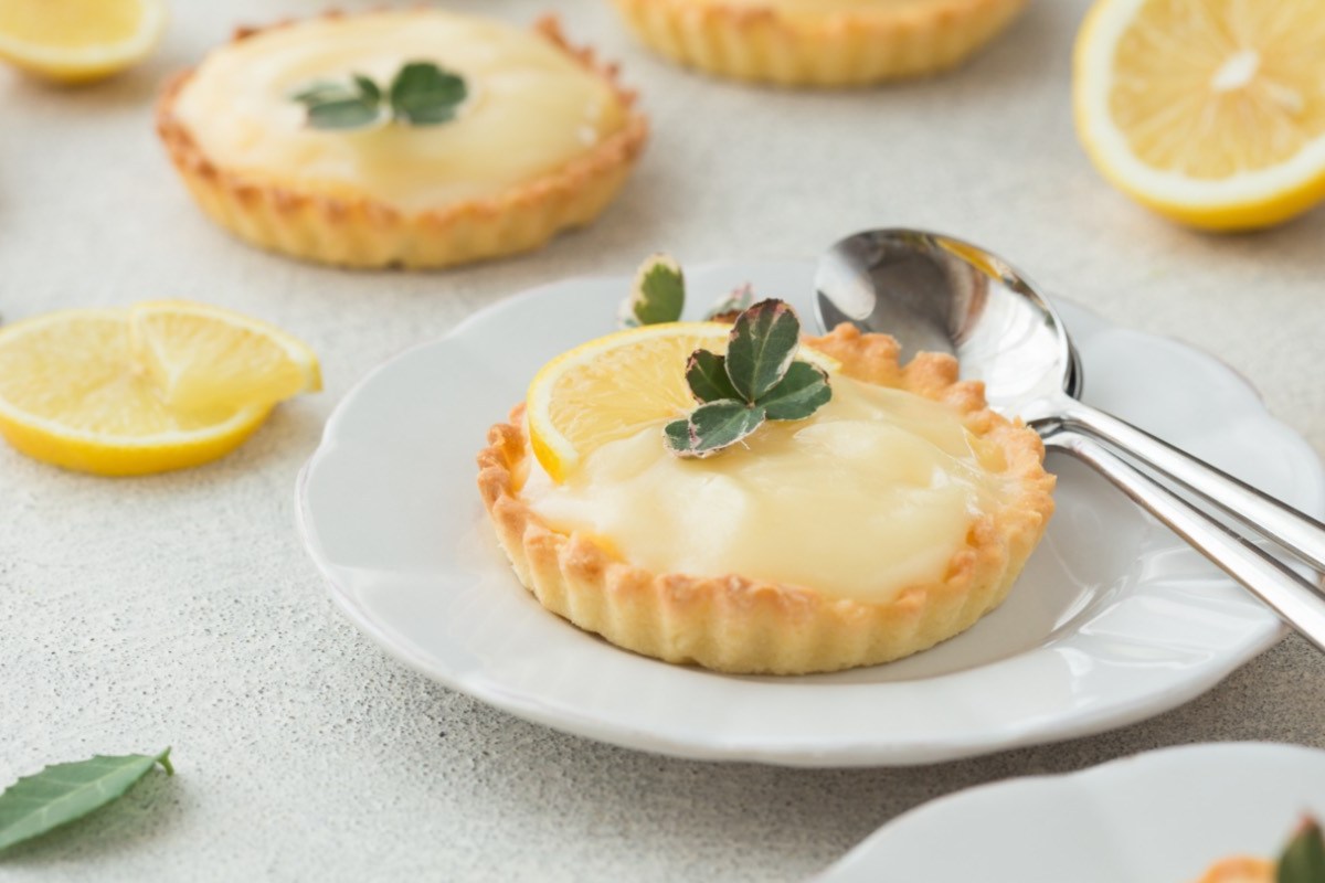 Zwei Lemon Curd Tartelettes auf Tellern. Neben einem liegt Besteck, im Hintergrund aufgeschnittene Zitronen.